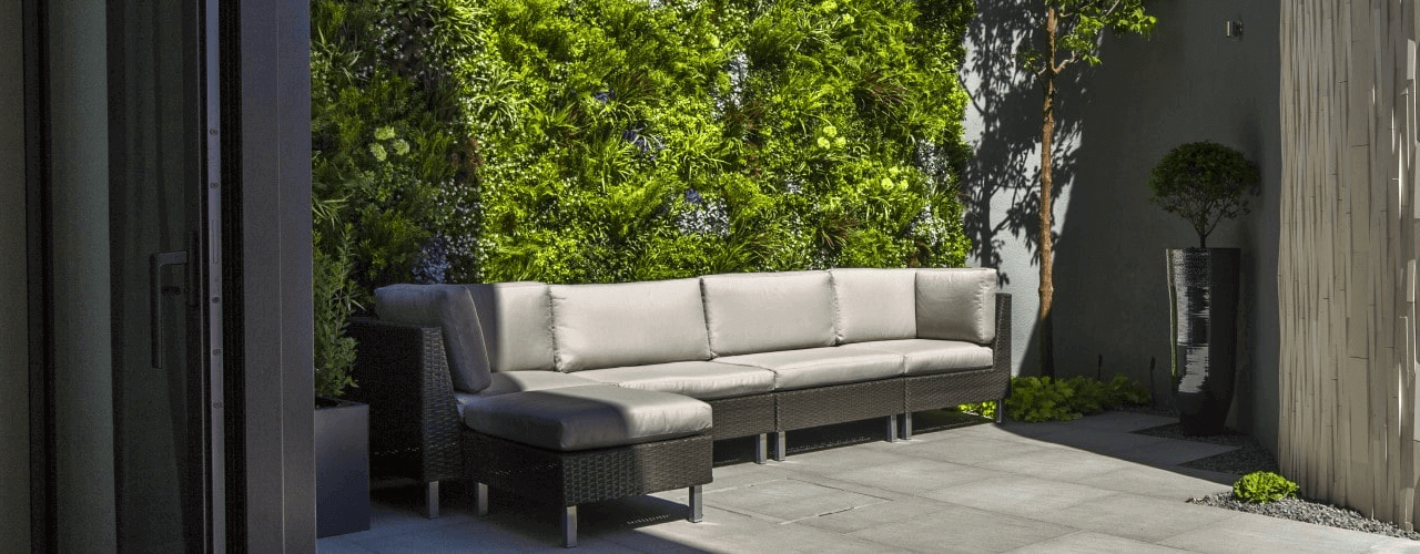 A modern outdoor patio features an artificial vertical garden wall behind a gray cushioned sectional sofa. The tiled floor complements the area, with a large potted plant next to the sofa, enhancing the greenery of this stylish space.