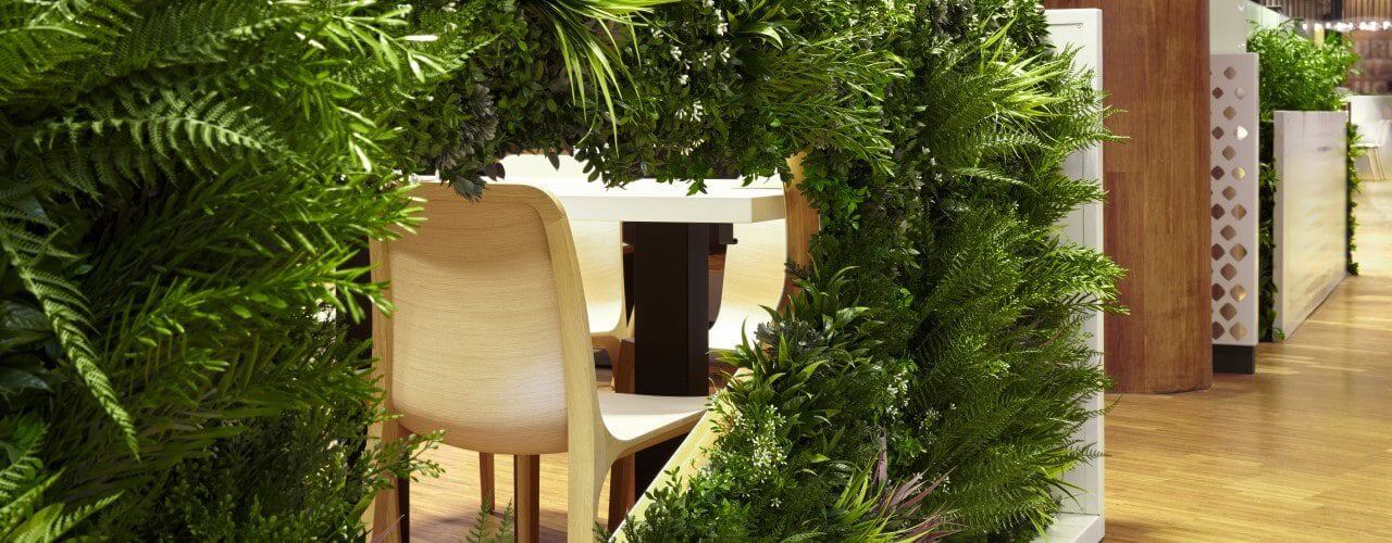 An indoor dining area with chairs and tables partially enclosed by lush, green foliage and plants. Privacy is enhanced by artificial living green vertical screen panels, complementing the natural wood tones to create a cozy, nature-inspired atmosphere.