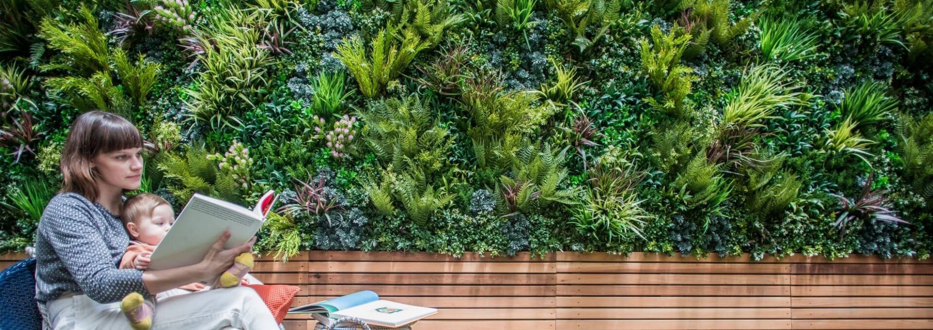 A woman reads a book with her baby in front of a lush green wall. They sit on a wooden bench, surrounded by books and papers. The vertical gardens foliage panels create a vibrant, natural setting, filled with various plants in the background.