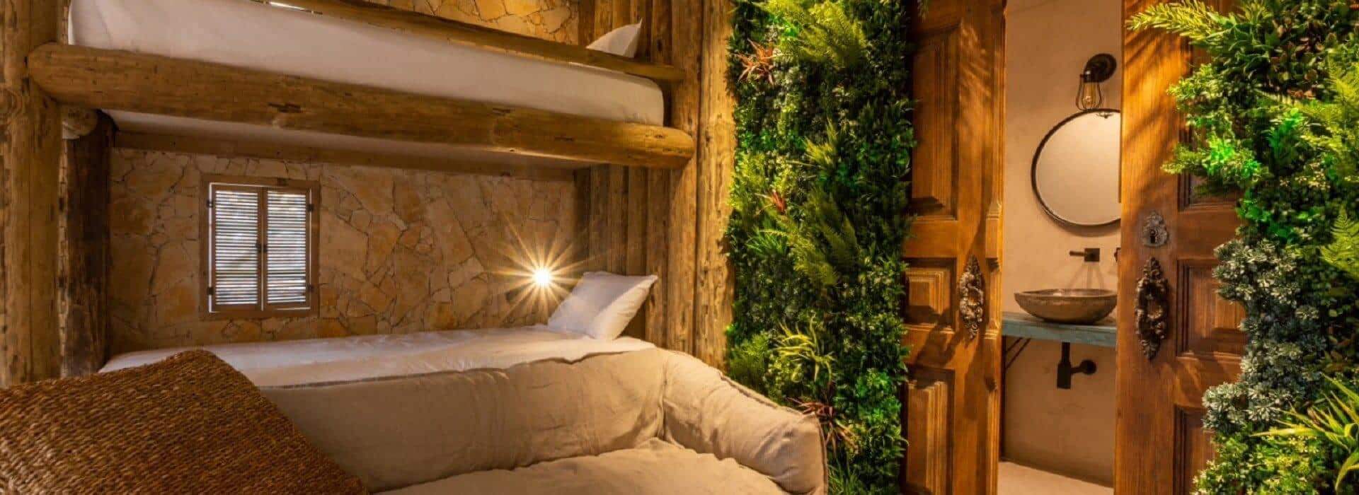 A cozy room featuring bunk beds with wooden accents and a small window overlooks a green wall for privacy screening. An open door reveals a rustic bathroom with a round mirror and stone basin sink. Warm lighting enhances the inviting atmosphere, reminiscent of the serene Azores.