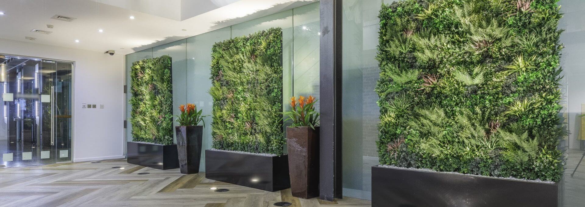 The modern lobby boasts a Mobile Green Wall Solution with three greenery walls and large plant pots with vibrant orange flowers. The space features wooden herringbone flooring, glass partitions, and spotlights, crafting a natural and contemporary atmosphere.