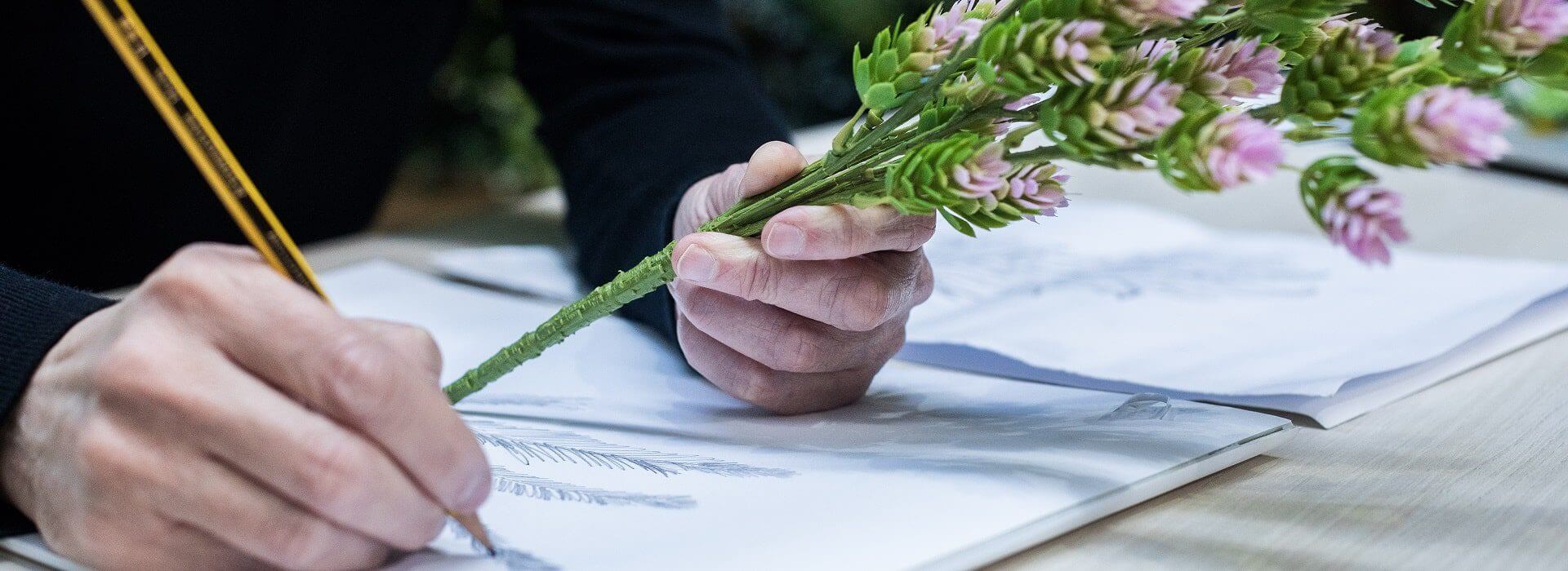 A person is sketching on paper while holding a stem of pink and green flowers, blending natural beauty with an artificial flair. The table is scattered with sheets as the pencil dances across the page, focusing on the intricate movements of the hands, the pencil, and the delicate flowers.