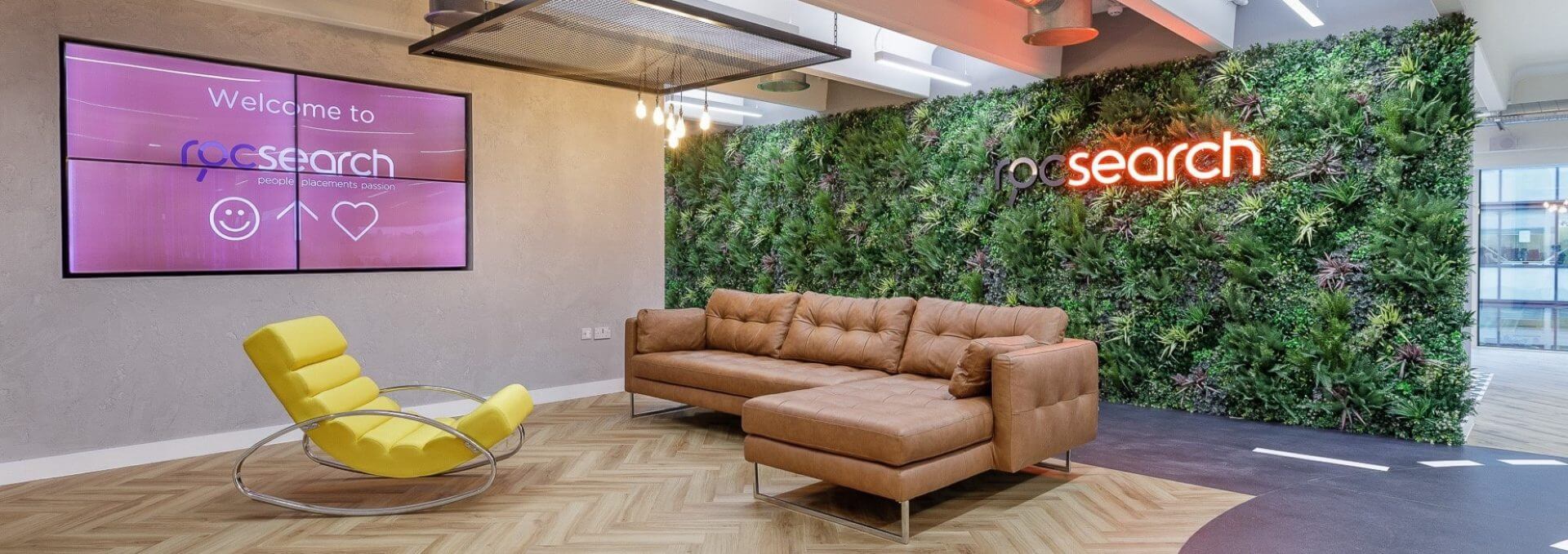 The modern office lobby in Reading, UK, features a brown leather L-shaped couch, a yellow rocking chair, and Roc Search's signature green wall with a neon sign that reads "pg search." Two screens on the opposite wall display a welcoming message.