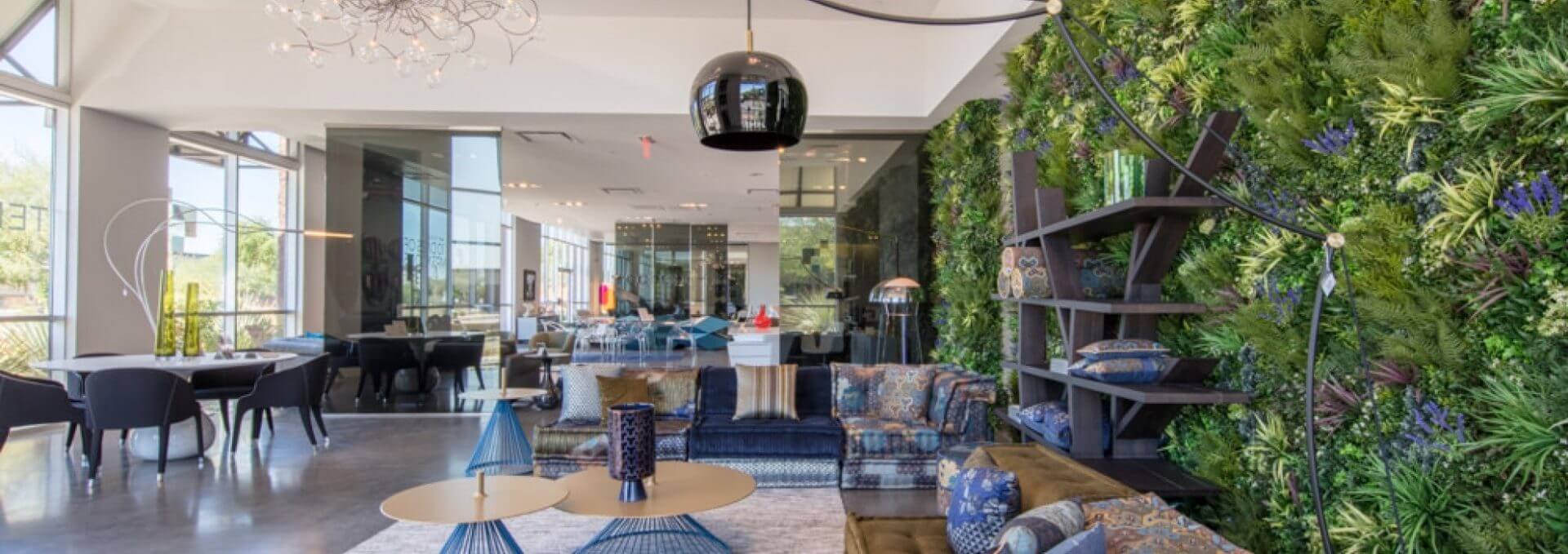 Modern spacious lobby with a lush green vertical garden on the wall, echoing the style of the Roche Bobois Showroom's Green Wall in Scottsdale. The room features stylish seating, round tables, and large windows allowing natural light to flood in. A black pendant light hangs from the ceiling, creating a cozy atmosphere.