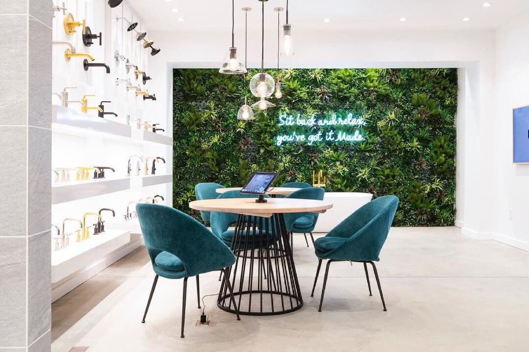 A modern showroom featuring a round table with teal chairs, a wall of various bathroom fixtures, and a lush green plant wall with a neon sign reading Sit back and relax, you’ve got it Made. Pendant lights hang from the ceiling.