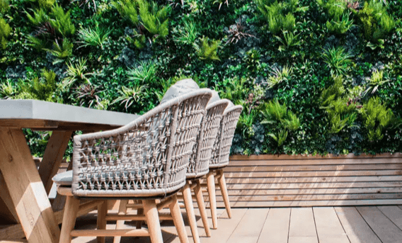 A modern outdoor seating area features a wooden table, wicker chairs, and an artificial vertical garden wall lush with various green plants. The wooden deck flooring and bench add to the natural ambiance of the tranquil space.