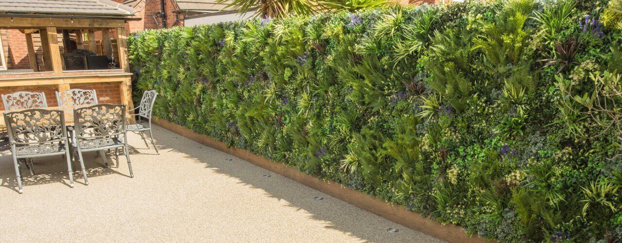 A sunny patio features a vertical garden wall lush with various green plants. To the left, decorative metal chairs sit near a wooden structure, possibly a gazebo or pergola. The ground is covered with light-colored tiles, enhanced by the vibrant addition of artificial elements for year-round beauty.