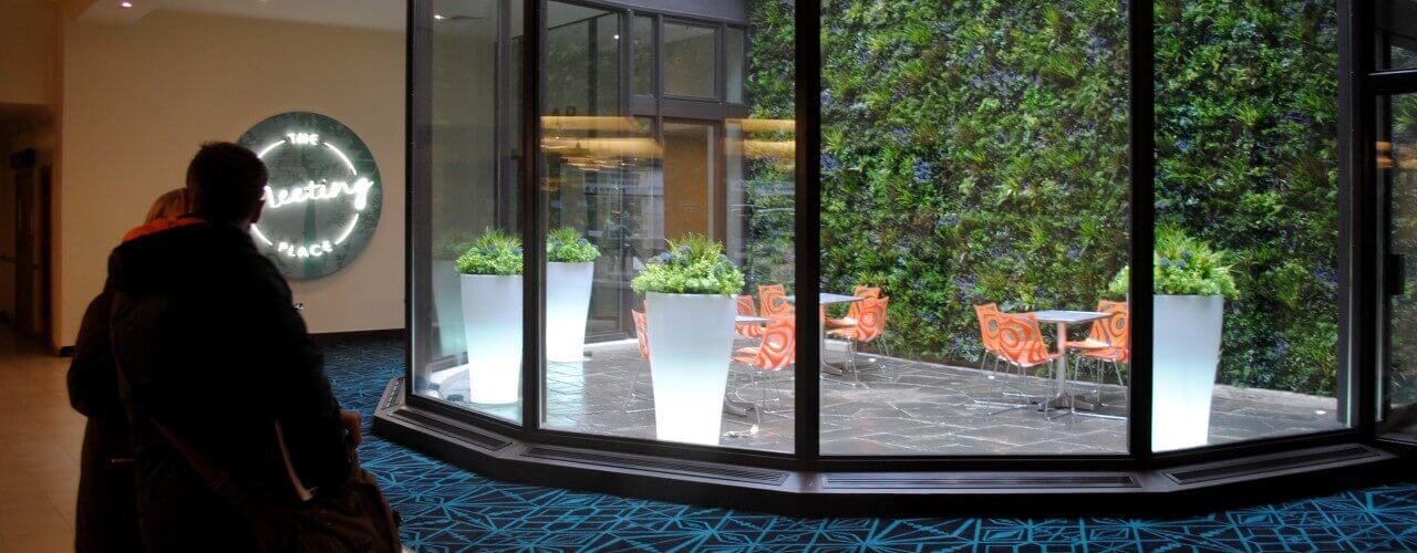 Two people peer through glass at a modern seating area with orange chairs and white tables. A green wall adorned with plants decorates the back, while a sign reads The Eating Place. Artificial blue geometric-patterned carpet adds to the vibrant atmosphere.