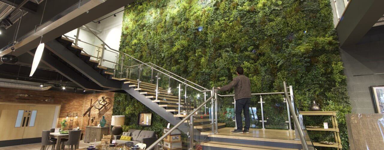 A person stands on a modern staircase admiring an artificial vertical garden wall. The room features industrial elements, contemporary furniture, and decorative pieces on display.