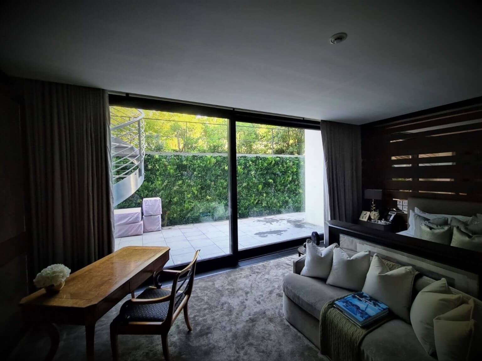 A modern room features a wooden desk and chair, a gray sofa with white pillows, and a large sliding glass door leading to a patio. Outside, an artificial spiral staircase and lush greenery invite ample natural light inside.