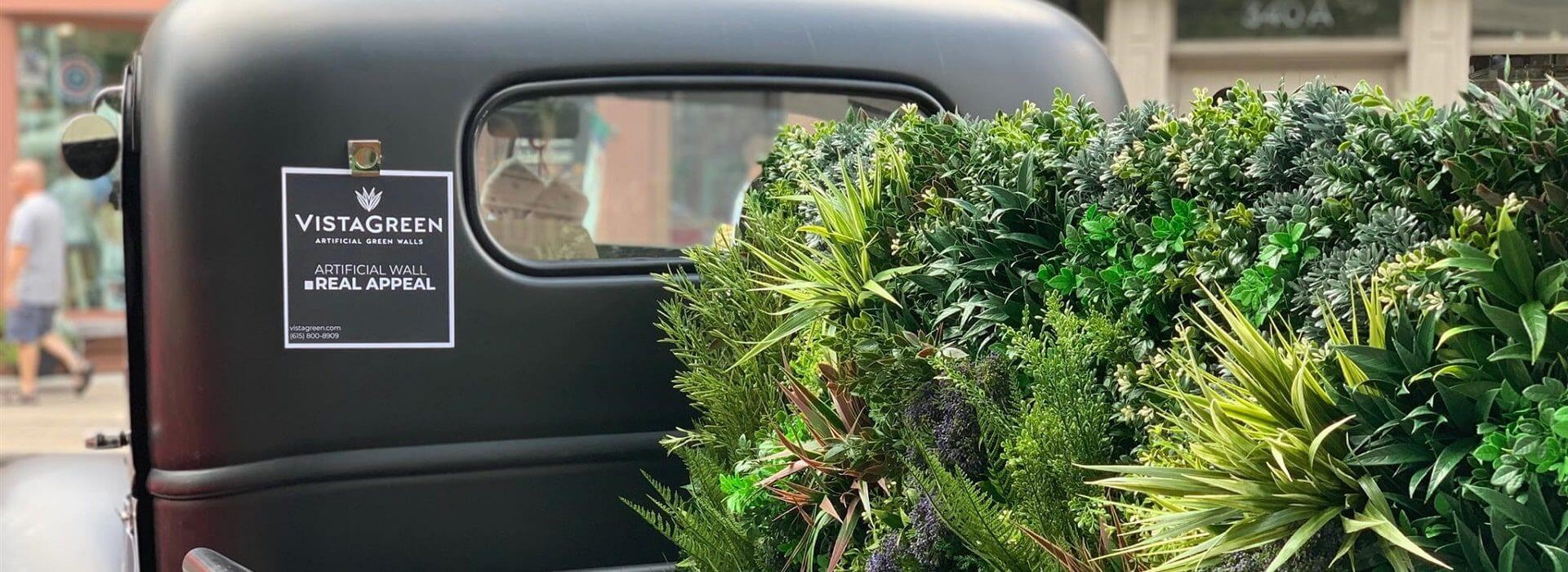 A classic car is adorned with a lush, artificial green wall in the back, echoing Fourth of July greenery foliage displays in Nashville. A sign reading "Vistagreen: Artificial Wall Real Appeal" graces the vehicle, adding charm against a backdrop of a shop and sidewalk reminiscent of Tennessee's vibrant streets.