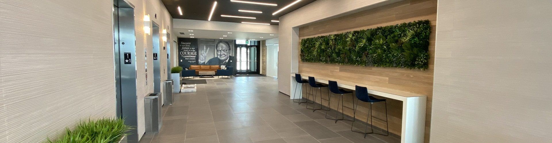 A modern office lobby in Lehi features a row of barstools and a vibrant green wall. The well-lit space includes sleek lighting, an elevator on the left, and a large mural with a quote and image on the far wall, embodying the innovative spirit of Utah's office spaces.