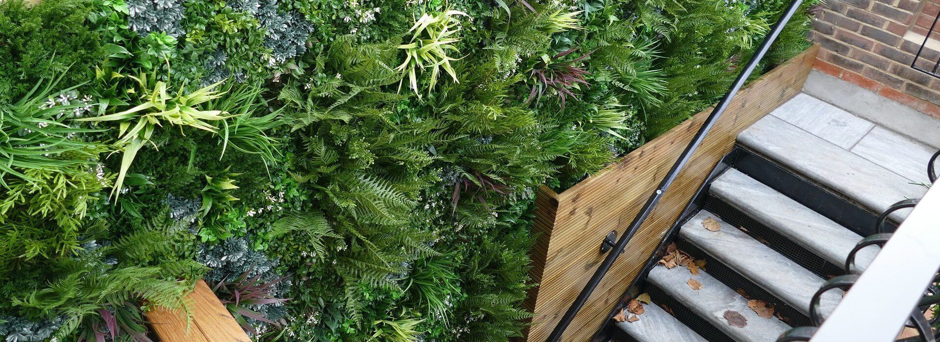 A lush artificial green wall filled with various plants covers a space next to the staircase. The stairs have wooden and metal railings, and a few brown leaves are scattered on the steps.