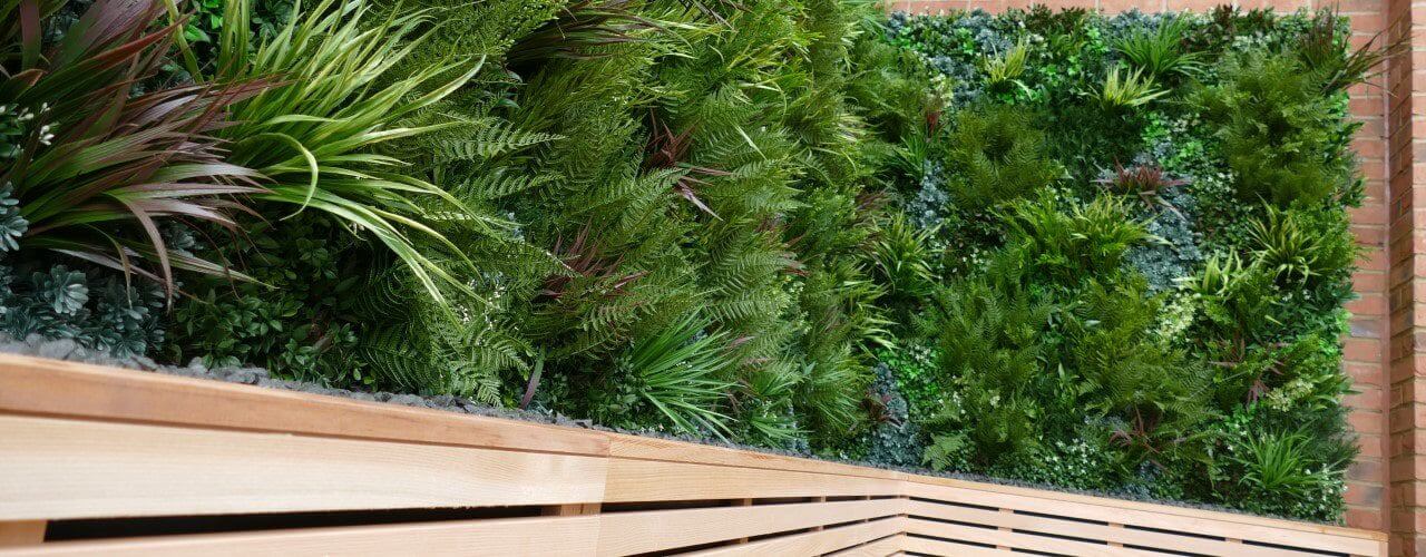 A vertical garden adorned with lush artificial plants and ferns blankets a brick wall. In the foreground, a wooden bench with a slatted design offers inviting seating adjacent to this vibrant display.