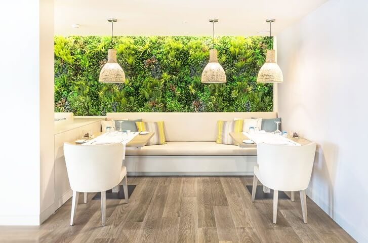 A modern dining area featuring Artificial Green Wall Panels, surrounded by white chairs and a light wood floor. The space includes a central cushioned bench with two tables decorated with tableware, complemented by three hanging pendant lights above.