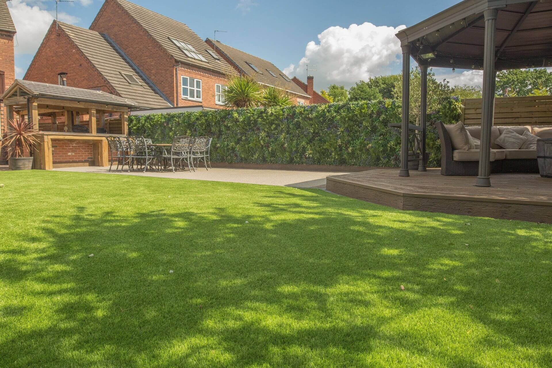 A green lawn with shadows covers the foreground, leading to a patio with a dining table and chairs. A brick barbecue area and a wooden gazebo are set against a backdrop of a vine-covered wall and red brick houses. The sky is partly cloudy.