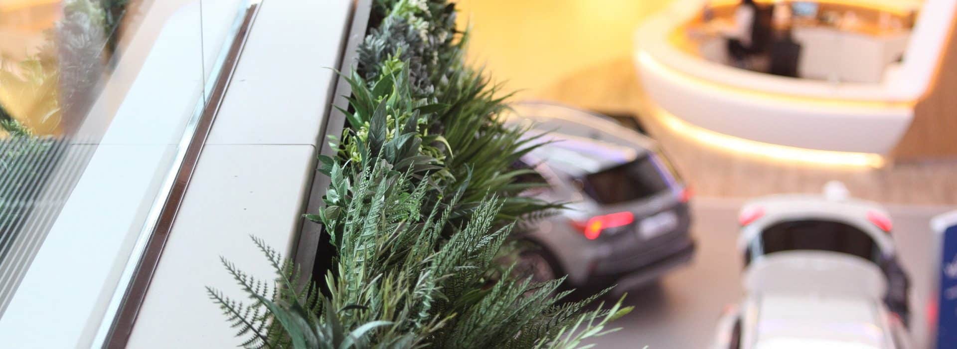 A sleek, modern interior with a focus on green plants beside a glass railing and an innovative Green Escalator in Sint-Niklaas. Below, blurred figures and two parked cars in a showroom setting add to the contemporary atmosphere.