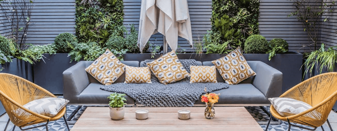 A stylish outdoor patio featuring a gray sofa adorned with patterned yellow and gray cushions. A gray throw is draped over the sofa. Two yellow chairs flank a wooden coffee table with candles and a flower vase. Lush greenery, thanks to vertical garden design, transforms your living space.