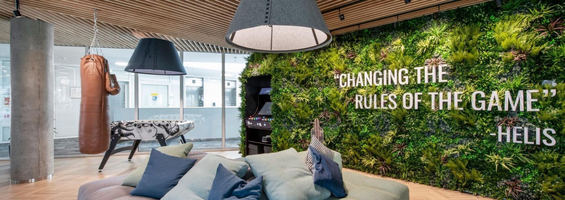 A modern office space in Kaunas, Lithuania, features a lush green wall with the quote "Changing the Rules of the Game" by Helis. Evoking workplace motivation, a plush sofa with pillows is in the foreground, while a punching bag and foosball table are visible in the background.