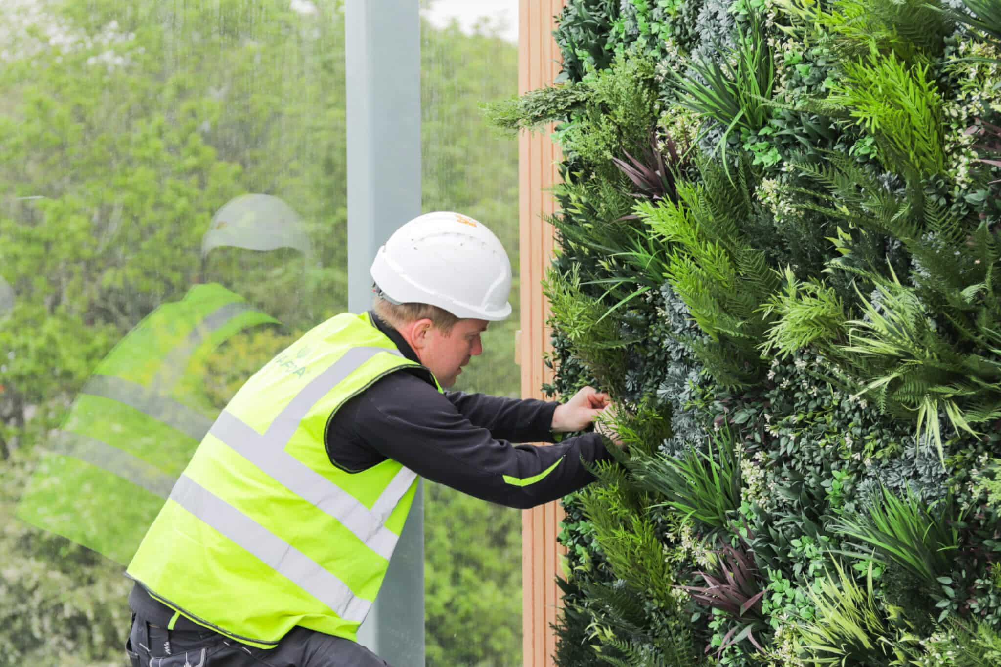 A worker in a neon yellow vest and white hard hat is tending to a vertical garden wall with lush green plants. A reflection is visible in a glass panel. In the background, there are trees and greenery.