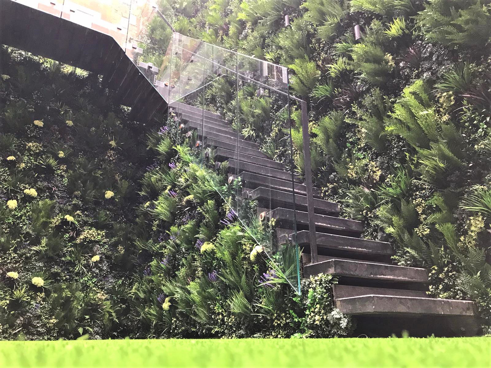 A modern staircase made of glass and metal ascends through a lush vertical garden wall, abundant with green artificial foliage and artificial yellow flowers.