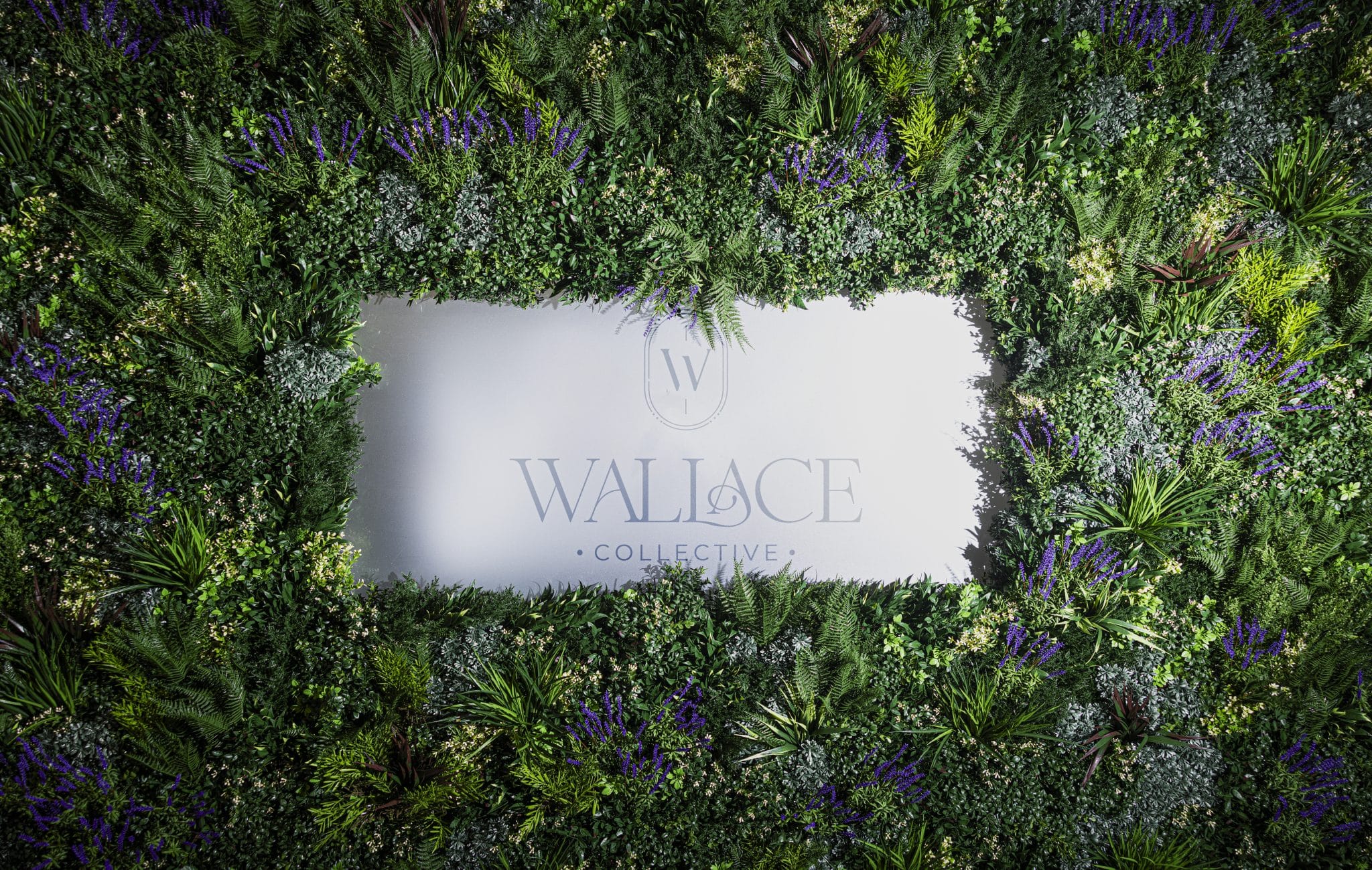 A rectangular sign with the text Wallace Collective is surrounded by a lush border of green plants and purple flowers.